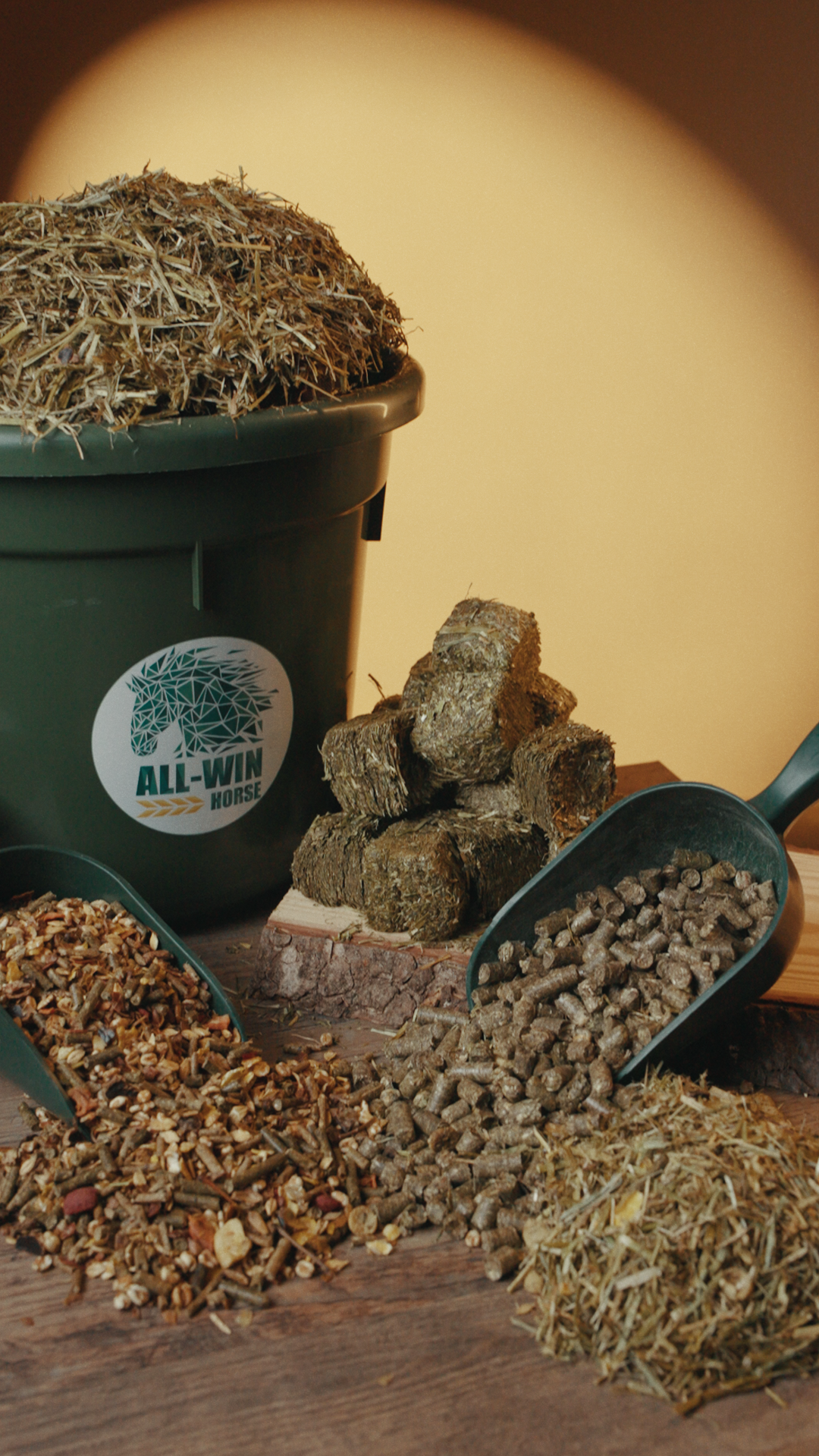 Grüner Futtersack mit All-Win-Horse-Logo, gefüllt mit losem Heu. Daneben liegen gepresste Heu-Quader, Pellets sowie verschiedene Müslimischungen in Futterschaufeln und kleinen Haufen dekorativ angerichtet auf Holz.